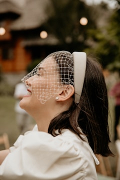 A woman with dark hair is wearing a white headband with an attached net veil, smiling and looking upwards. She is dressed in a white outfit with pearl earrings visible. The background is blurred, featuring greenery and what appears to be wooden structures, suggesting an outdoor setting.