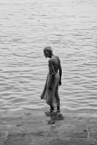 An elderly man standing barefoot on a quiet, sunlit beach, completely natural and unadorned.