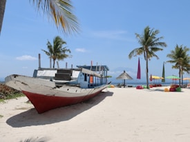 A wooden boat with a red hull rests on a sandy beach beside vibrant, multicolored umbrellas and cushioned seating. The sky is clear and blue, and palm trees are scattered around, providing a tropical atmosphere.