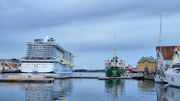 A large cruise ship and a smaller green ship are docked at a harbor. The cruise ship is prominently positioned on the left, with a white and blue color scheme. The green ship with a tall mast is docked closer to the right. Surrounding buildings include houses with red-tiled roofs and a yellow house with distinct architecture. The water is calm, reflecting the ships and buildings. The sky is overcast with light clouds.