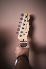 A hand holding the neck of a Squier Telecaster guitar in a relaxed grip. The background is a muted brown wall, providing a neutral backdrop to the light-colored wood and metal tuners of the guitar headstock.