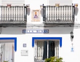 A white building exterior with two decorative iron balconies and colorful flower pots. The sign reads 'Villa Susan' in blue. Below is a door and a number plate labeled 37. A small religious tile image and a cross are on the wall, along with a black lantern.