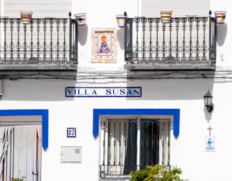 A white building exterior with two decorative iron balconies and colorful flower pots. The sign reads 'Villa Susan' in blue. Below is a door and a number plate labeled 37. A small religious tile image and a cross are on the wall, along with a black lantern.