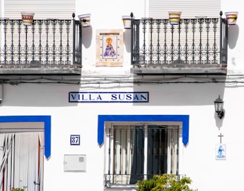 A white building exterior with two decorative iron balconies and colorful flower pots. The sign reads 'Villa Susan' in blue. Below is a door and a number plate labeled 37. A small religious tile image and a cross are on the wall, along with a black lantern.