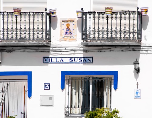 A white building exterior with two decorative iron balconies and colorful flower pots. The sign reads 'Villa Susan' in blue. Below is a door and a number plate labeled 37. A small religious tile image and a cross are on the wall, along with a black lantern.