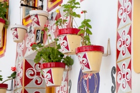 Multiple decorative flower pots with red and white geometric patterns are mounted on a vibrantly painted wall. The wall features intricate designs consisting of swirling shapes and diamond patterns in red, white, and gold. Green plants can be seen flourishing from the pots, adding a touch of nature to the structured and colorful setup.
