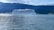 A large white cruise ship sails on calm waters near a mountainous landscape. The ship has multiple decks and distinctive blue wave patterns on its sides. The surrounding scenery includes lush green hills and a partly cloudy sky.