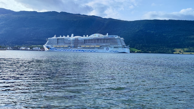 A large white cruise ship sails on calm waters near a mountainous landscape. The ship has multiple decks and distinctive blue wave patterns on its sides. The surrounding scenery includes lush green hills and a partly cloudy sky.