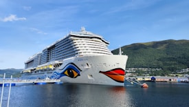 A large cruise ship is docked at a pier in a scenic coastal area. The ship is white with colorful decorations on its hull, including a prominent eye and wave patterns. The coastline features green hills and a small town with buildings along the shore. The sky is clear with minimal clouds.