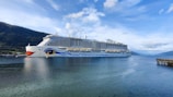 A large cruise ship with a distinctive eye and wave design on its hull navigates through a calm body of water. Surrounding the ship are scenic, tree-covered hills and a clear sky with scattered clouds. To the right, a pier extends slightly into the water.