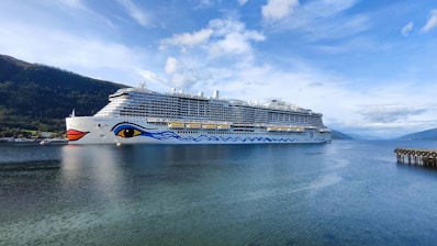 A large cruise ship with a distinctive eye and wave design on its hull navigates through a calm body of water. Surrounding the ship are scenic, tree-covered hills and a clear sky with scattered clouds. To the right, a pier extends slightly into the water.