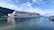 A large cruise ship with a distinctive eye and wave design on its hull navigates through a calm body of water. Surrounding the ship are scenic, tree-covered hills and a clear sky with scattered clouds. To the right, a pier extends slightly into the water.
