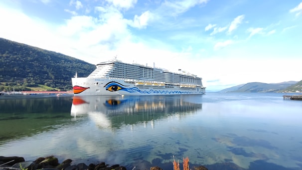 A large cruise ship with distinctive eye-like designs on the side sails through a calm body of water, reflecting its image perfectly. Surrounding the vessel are serene mountain landscapes under a clear blue sky.