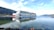 A large cruise ship with distinctive eye-like designs on the side sails through a calm body of water, reflecting its image perfectly. Surrounding the vessel are serene mountain landscapes under a clear blue sky.