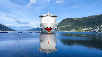 A large cruise ship is docked in a calm, scenic fjord with mountains in the background. The ship has a distinctive painted design resembling a smiling face with lips at the bow. The clear water reflects the ship and the sky, contributing to a serene atmosphere.