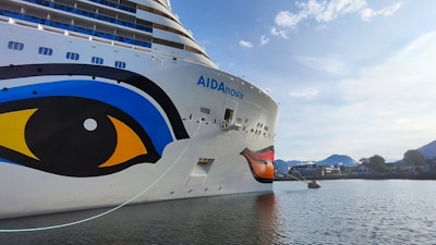 The image features a large cruise ship with a distinctive eye and lip design painted on its hull. The ship is named AIDAnova and is docked at a port. In the background, there are calm waters and a mountainous landscape under a partly cloudy sky.