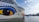 The image features a large cruise ship with a distinctive eye and lip design painted on its hull. The ship is named AIDAnova and is docked at a port. In the background, there are calm waters and a mountainous landscape under a partly cloudy sky.