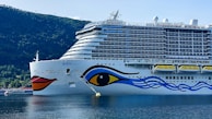 A large cruise ship with a colorful design on its hull, featuring an eye and abstract blue and red patterns. The ship is docked in a calm body of water, surrounded by lush green hills under a clear blue sky.