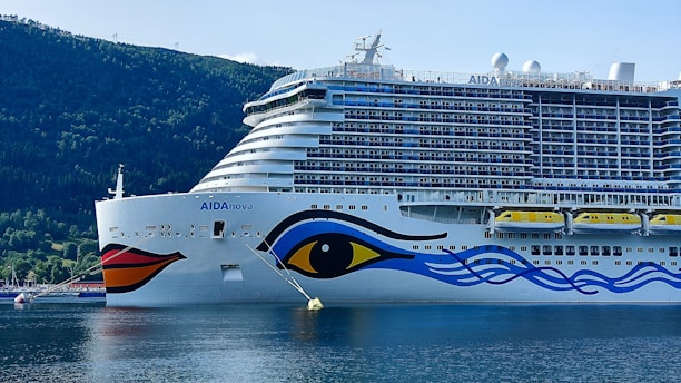 A large cruise ship with a colorful design on its hull, featuring an eye and abstract blue and red patterns. The ship is docked in a calm body of water, surrounded by lush green hills under a clear blue sky.