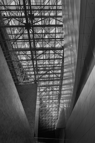 A complex lattice of steel beams forms the ceiling of a modern architectural space, casting intricate shadows on the walls. The space is defined by angular, geometric shapes and a high ceiling, creating a sense of openness.