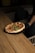 A freshly baked pizza with a thick crust and toppings of basil leaves sits on a round black tray. The pizza is being served by a person wearing a black glove, and it is placed on a wooden table with a warm, natural finish. The background features a dark, cozy setting with a glimpse of cushioned seating.