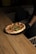 A freshly baked pizza with a thick crust and toppings of basil leaves sits on a round black tray. The pizza is being served by a person wearing a black glove, and it is placed on a wooden table with a warm, natural finish. The background features a dark, cozy setting with a glimpse of cushioned seating.