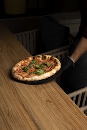 A freshly baked pizza with a thick crust and toppings of basil leaves sits on a round black tray. The pizza is being served by a person wearing a black glove, and it is placed on a wooden table with a warm, natural finish. The background features a dark, cozy setting with a glimpse of cushioned seating.
