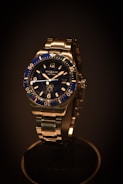 A luxury wristwatch with a metallic strap and a deep blue bezel. The watch face features clear hour markers and minute indices, with the brand name visible on the dial. The lighting emphasizes the sleek design and shiny surfaces.