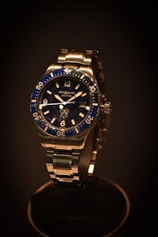 A luxury wristwatch with a metallic strap and a deep blue bezel. The watch face features clear hour markers and minute indices, with the brand name visible on the dial. The lighting emphasizes the sleek design and shiny surfaces.