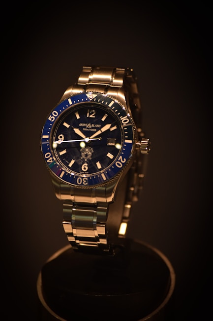 A luxury wristwatch with a metallic strap and a deep blue bezel. The watch face features clear hour markers and minute indices, with the brand name visible on the dial. The lighting emphasizes the sleek design and shiny surfaces.