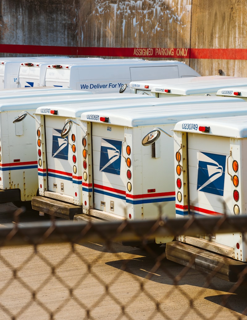 A row of US Postal Service mail trucks behind a chain link fence