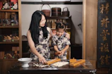 A woman and a young girl are engaged in a cooking activity at a wooden table, surrounded by traditional decorations and shelves with boxed items. Both are wearing matching floral-patterned dresses, and the woman is gently guiding the girl as they work with a wooden mold and dough. Warm lighting enhances the cozy, familial atmosphere.