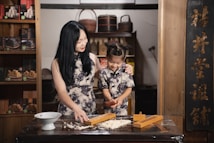 A woman and a young girl are engaged in a cooking activity at a wooden table, surrounded by traditional decorations and shelves with boxed items. Both are wearing matching floral-patterned dresses, and the woman is gently guiding the girl as they work with a wooden mold and dough. Warm lighting enhances the cozy, familial atmosphere.