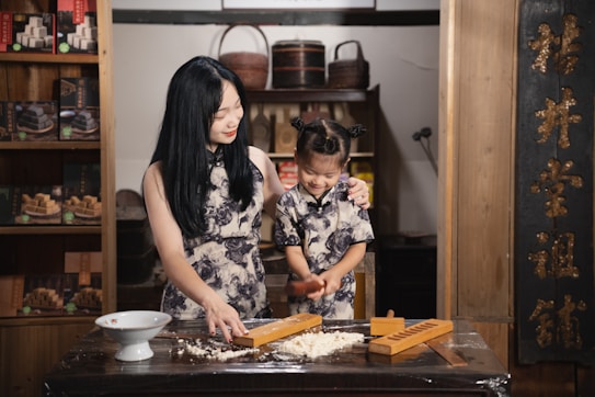 A woman and a young girl are engaged in a cooking activity at a wooden table, surrounded by traditional decorations and shelves with boxed items. Both are wearing matching floral-patterned dresses, and the woman is gently guiding the girl as they work with a wooden mold and dough. Warm lighting enhances the cozy, familial atmosphere.