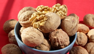 A blue bowl filled with whole walnuts, with one walnut cracked open to reveal its interior. The background is a vibrant red, contrasting with the natural brown tones of the walnuts.