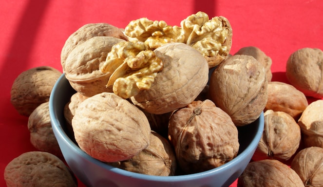 A blue bowl filled with whole walnuts, with one walnut cracked open to reveal its interior. The background is a vibrant red, contrasting with the natural brown tones of the walnuts.