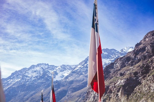 A flagpole with a multicolored flag is prominently displayed in front of a majestic mountain range. The mountains are covered with snow, and the sky is clear with a few scattered clouds.
