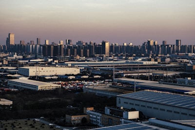 An expansive industrial area with large warehouses and factories stretches across the foreground, while a dense urban skyline with numerous high-rise buildings rises in the background under a dusky sky.