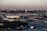 An expansive industrial area with large warehouses and factories stretches across the foreground, while a dense urban skyline with numerous high-rise buildings rises in the background under a dusky sky.