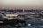 An expansive industrial area with large warehouses and factories stretches across the foreground, while a dense urban skyline with numerous high-rise buildings rises in the background under a dusky sky.