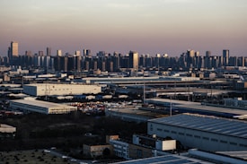An expansive industrial area with large warehouses and factories stretches across the foreground, while a dense urban skyline with numerous high-rise buildings rises in the background under a dusky sky.