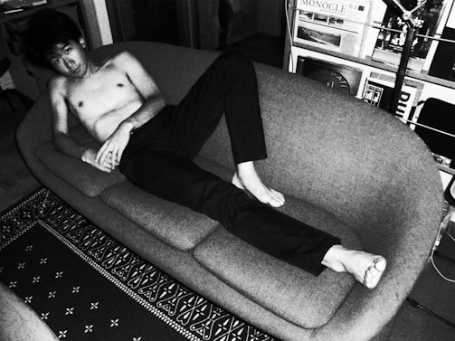 A person is lounging shirtless on a modern, curved sofa in a room filled with various magazines and a floor lamp. The setting appears to be an indoor living space with decorative elements such as a patterned rug on the floor. The image is in black and white, giving it a classic or vintage feel.
