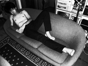 A person is lounging shirtless on a modern, curved sofa in a room filled with various magazines and a floor lamp. The setting appears to be an indoor living space with decorative elements such as a patterned rug on the floor. The image is in black and white, giving it a classic or vintage feel.