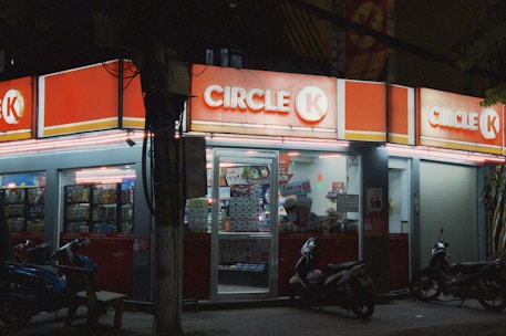 A convenience store is brightly lit and open at night, with a red and white color scheme. The entrance has glass doors displaying promotional posters. Several motorbikes are parked in front of the store.