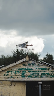 A weathered and peeling roof of a small structure featuring a rusty metal weather vane on top, set against a backdrop of cloudy skies. The roof has faded green and yellow paint, giving it an aged appearance.