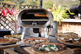 A modern outdoor pizza oven is surrounded by several pizzas, each with various toppings. The setting includes a wooden table, vibrant green plants, and a brick wall in the background, suggesting a cozy garden ambiance.