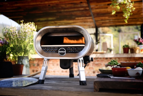 A modern outdoor pizza oven is set on a wooden table under a rustic pergola. The oven has a sleek stainless steel design, and the brand name 'Revolve' is visible on its front. Surrounding the oven are various cooking ingredients and plates with fresh vegetables and herbs, adding a vibrant touch. The background features potted plants and hanging greenery, enhancing the natural setting.
