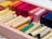A wooden box filled with an assortment of colorful oil pastels and crayons. The crayons are neatly arranged in rows with hues ranging from yellow, orange, red, to green and dark blue. There are also rectangular sticks of different colors beside the crayons.