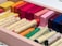 A wooden box filled with an assortment of colorful oil pastels and crayons. The crayons are neatly arranged in rows with hues ranging from yellow, orange, red, to green and dark blue. There are also rectangular sticks of different colors beside the crayons.