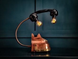 A vintage style lamp designed to resemble an old-fashioned telephone, with a copper base and a handset serving as the lamp arm. Two light bulbs hang from the handset, casting a warm glow. Set against a dark, teal background, the overall design blends retro and industrial elements.